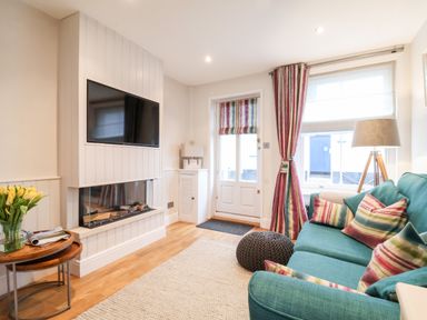 A living room with a teal sofa and striped cushions near a window with matching curtains and a lamp at 51 Tern Cottage Aldeburgh