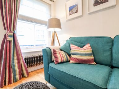 A corner of a living room with a teal sofa with striped cushions a window with striped curtains a floor lamp and framed pictures on the wall at 51 Tern Cottage Aldeburgh