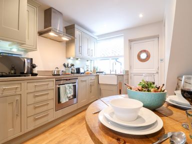 A kitchen with beige cabinets and countertops a round wooden dining table set with plates bowls salad and wine glasses at 51 Tern Cottage Aldeburgh