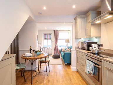 A kitchen and dining area with a wooden table set for two leading to a living area with a blue sofa and pouf at 51 Tern Cottage Aldeburgh