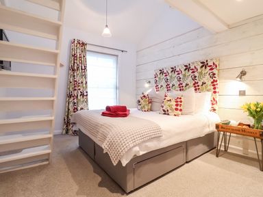 A bedroom with a double bed with floral pillows and headboard a side table with flowers and a staircase at 51 Tern Cottage Aldeburgh