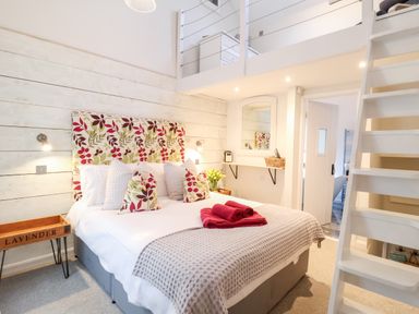 A bedroom with a bed with floral pillows and folded towels with a loft area above and a staircase at 51 Tern Cottage Aldeburgh
