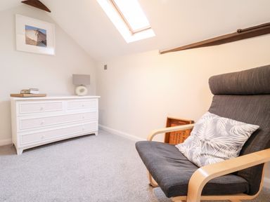 A room with a white dresser with books and a lamp and a gray armchair with a patterned cushion at 51 Tern Cottage Aldeburgh