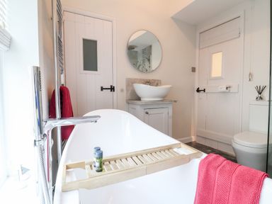 A bathroom with a freestanding bathtub a towel on the side a wooden bath tray with toiletries a circular mirror above a sink and a toilet at 51 Tern Cottage Aldeburgh