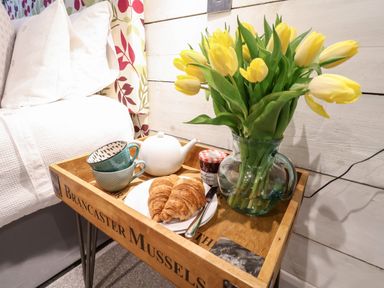 A wooden tray table with yellow tulips in a glass pitcher two teacups a teapot croissants and jam next to a bed in a bedroom at 51 Tern Cottage Aldeburgh