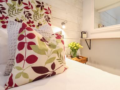 A bed with pillows featuring red and green leaf patterns next to a wall with a lamp a mirror and a tray holding yellow flowers and a radio at 51 Tern Cottage Aldeburgh