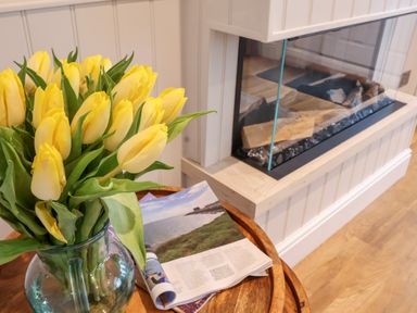 A bouquet of yellow tulips in a glass vase on a wooden table next to a magazine by a glass-enclosed fireplace at 51 Tern Cottage Aldeburgh