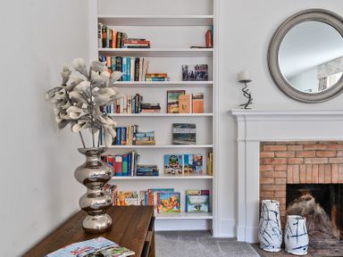 A bookshelf with books and a wooden table with a metallic vase next to a fireplace with blue and white ceramic vases and a round mirror on the wall at Bedingfield House nr Debenham Debenham