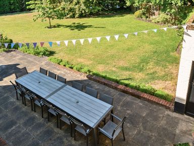 An outdoor patio with a long rectangular table and chairs on a paved area overlooking a lawn with a tree and bunting at Bedingfield House nr Debenham Debenham
