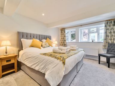 A bedroom with a grey upholstered bed with white bedding and yellow pillows beside wooden nightstands with lamps and a grey chair near windows with patterned curtains at Bedingfield House nr Debenham Debenham