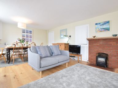 A living room with a gray sofa blue cushions a dining table with chairs flowers on the table a brick fireplace and a television at Seaflower Aldeburgh