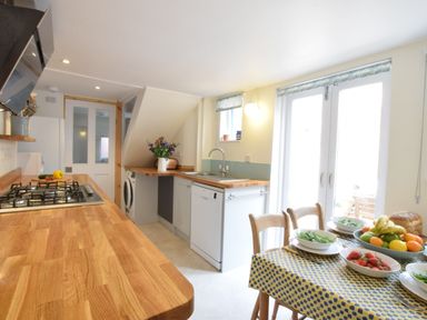 A kitchen with wooden countertops a gas stove a dishwasher washing machine a dining table with bowls of fruit and salad and double doors at Seaflower Aldeburgh