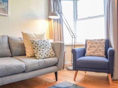 A living room with a grey sofa and a blue armchair with cushions and a floor lamp by the window at Seaflower in Aldeburgh