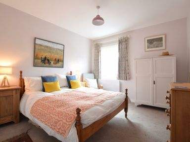 A bedroom with a wooden bed with pillows and a blanket a wooden nightstand with a lamp a white wardrobe a dresser an armchair and a window with curtains at Seaflower Aldeburgh in Aldeburgh