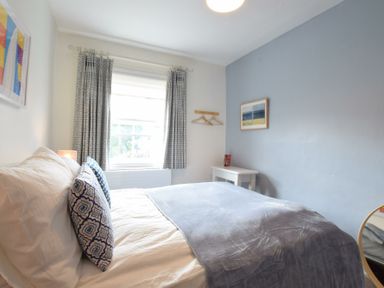 A bedroom with a bed, two patterned pillows, a window with curtains, a small white table, two wall hangers, and framed artwork at Seaflower, Aldeburgh