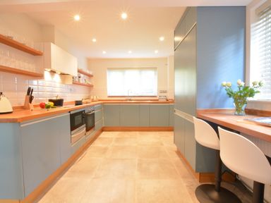 A kitchen with blue cabinets wooden countertops a sink under a window and two white chairs at a breakfast bar at Town Farm House Orford