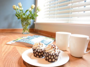 A kitchen counter with two cupcakes on a plate two white mugs a vase of white roses and magazines near a window with blinds at Town Farm House Orford