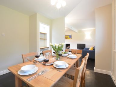 A dining table set with plates, bowls, wine glasses, bread, and a vase of flowers in a room with a living area and a television in the background at Town Farm House, Orford