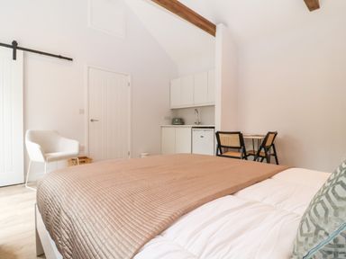 A bedroom with a bed covered by a quilt a white armchair a small kitchenette and a table with two chairs at Town Farm House Orford