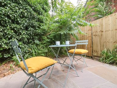 An outdoor patio with two metal chairs with yellow cushions and a small round table with two mugs surrounded by greenery and wooden fencing at Town Farm House Orford