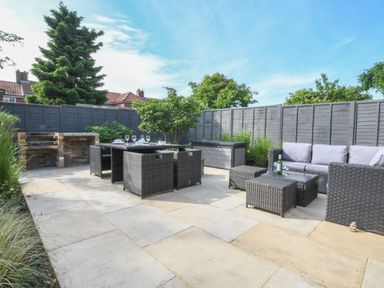 An outdoor patio with wicker dining set and seating area next to a brick barbecue and gray wooden fence at Corner Cottage Thorpeness