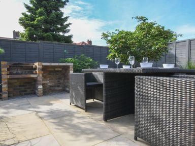 An outdoor patio with wicker dining furniture set and a built in brick barbecue with plants and fence in the background at Corner Cottage Thorpeness