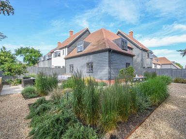 A garden with gravel paths and various plants around a gray wooden house with a tiled roof at Corner Cottage Thorpeness