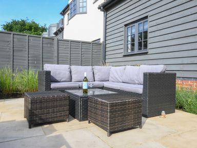 An outdoor patio with a gray cushioned sectional sofa and wicker furniture including a glass top table and two ottomans at Corner Cottage Thorpeness