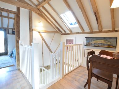 An upstairs hallway with wooden beams a skylight white railing a wooden desk and a bedroom visible at Fishpond House Sotherton in Halesworth