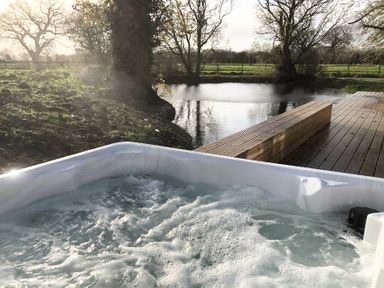 A bubbling hot tub next to a pond with trees and a wooden deck at Fishpond House Sotherton in Halesworth