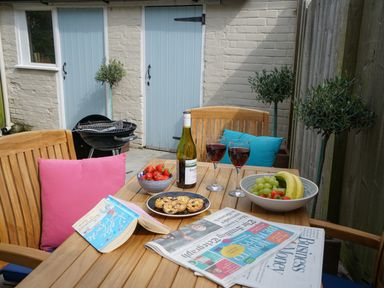 A wooden outdoor table with fruit bowls, cookies, wine glasses, a book, and newspapers on a patio at Smugglers Rest Aldeburgh