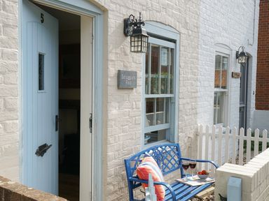 A blue bench with pillows and glasses of wine outside a white brick house with a blue door and windows at Smugglers Rest Aldeburgh