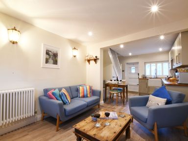 A living room with blue sofas colorful cushions and a wooden coffee table next to a dining area and kitchen at Smugglers Rest Aldeburgh