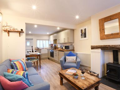 A living room with a blue sofa and armchair a wooden coffee table with two glasses of wine and a kitchen with cabinets and appliances at Smugglers Rest Aldeburgh