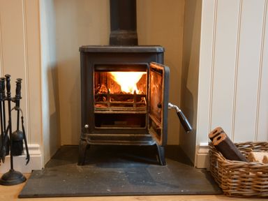 A wood burning stove with fire inside a corner of a room with fireplace tools and a basket of firewood at Smugglers Rest Aldeburgh