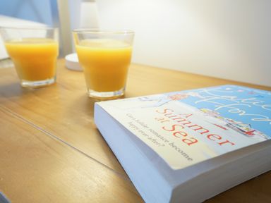 A wooden table with two glasses of orange juice and a book titled A Summer at Sea at Smugglers Rest in Aldeburgh