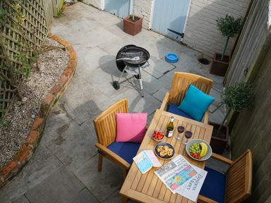 A patio with wooden table and chairs with cushions a barbecue and plants at Smugglers Rest Aldeburgh