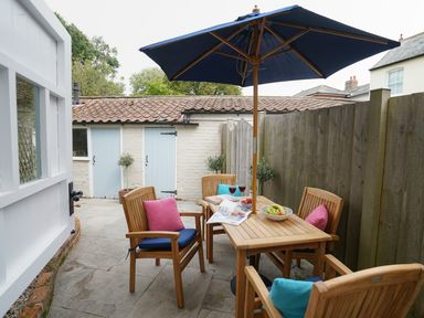 An outdoor patio with a wooden table and chairs under a blue umbrella with cushions and fruit bowl at Smugglers Rest Aldeburgh