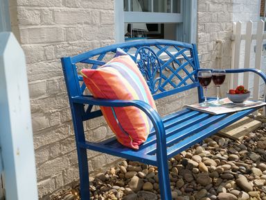 A blue metal bench with a colorful striped pillow two glasses of red wine a bowl of strawberries and a newspaper on a pebble patio at Smugglers Rest Aldeburgh