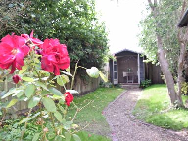 A garden with red flowers in the foreground a gravel path leading to a wooden shed with open doors and a small table and chair inside at Rosemary Cottage Aldeburgh