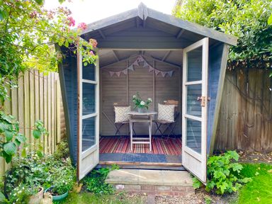 A wooden garden shed with open glass doors showing a small table with two chairs and cushions a potted plant on the table bunting on the back wall and a striped rug on the floor at Rosemary Cottage Aldeburgh