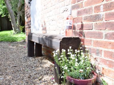 A wooden bench against a brick wall with a bottle of wine and glass on top and a flower pot with white flowers next to it in a garden