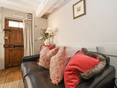 A living room with a black sofa and cushions near a wooden door and a small cabinet at Rosemary Cottage Aldeburgh