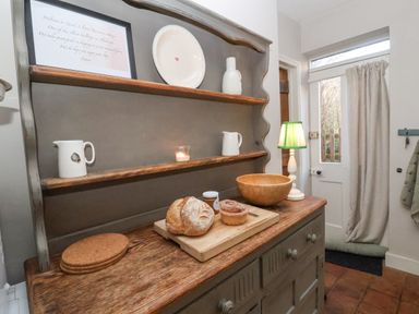 A wooden dresser with shelves holding a framed note, plates, jugs, a lit candle, bread, a pastry, a jar, a wooden bowl, and a lamp near a door with a curtain at Rosemary Cottage Aldeburgh