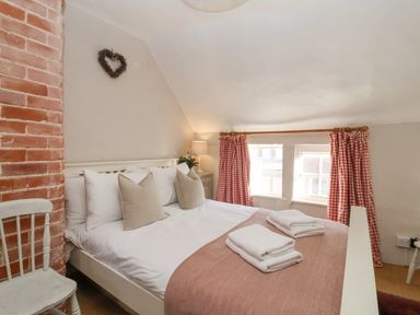 A bedroom with a white bed and pillows two windows with red check curtains and a wooden chair at Rosemary Cottage in Aldeburgh