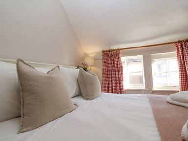 A bedroom with a bed with beige pillows white sheets a folded towel lamp and red checkered curtains at Rosemary Cottage Aldeburgh