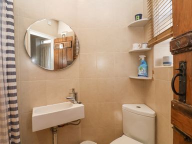 A small bathroom with a wall-mounted sink and round mirror above a toilet with corner shelves holding cups and a cleaning spray at Rosemary Cottage Aldeburgh