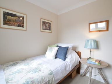 A single bed with pillows and a floral throw next to a wooden side table with a lamp and books at Napier House Southwold