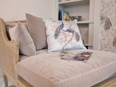 A beige cushioned wooden chair with pillows including one with peacock design and a magazine on seat at Napier House Southwold