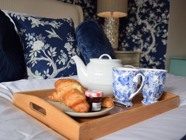 A breakfast tray with croissants jam a white teapot and two blue and white mugs on a bed with blue floral cushions at Napier House Southwold